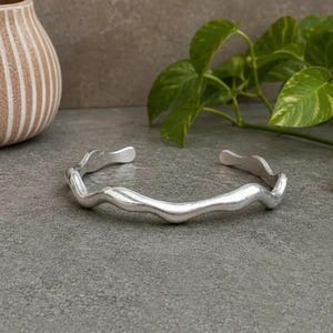 May include: A silver-toned cuff bracelet featuring a continuous wavy, undulating design. The metal has a smooth, polished finish with rounded edges, forming an open-ended circular shape that curves gently along its entire length.