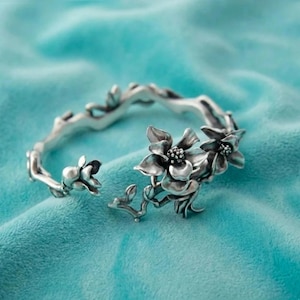 Puede incluir: Una pulsera plateada con un diseño floral. La pulsera presenta flores y tallos detallados, con un diseño abierto. La joya está sobre un fondo turquesa.