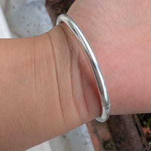 Może przedstawiać: Błyszcząca, srebrna bransoletka typu bangle. Bransoletka ma prosty, zaokrąglony kształt i wydaje się być wykonana z metalu. Bransoletka jest noszona na nadgarstku. Bransoletka ma średnicę około 6,35 cm.