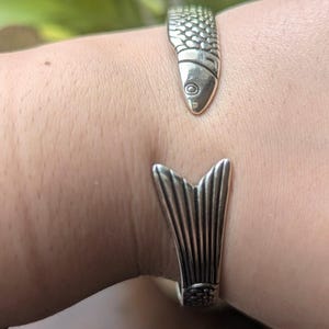 May include: A silver-toned open cuff bracelet shaped like a fish. One end of the bracelet features a detailed fish head with embossed scales, while the other end displays a wide, fan-shaped tail fin with vertical grooves and a textured base.