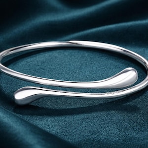 May include: A silver-coloured, open cuff bracelet with a teardrop design at each end. The bracelet is displayed on a dark teal fabric, creating a contrast that highlights the jewellery's shine. The bracelet's design is simple and elegant.
