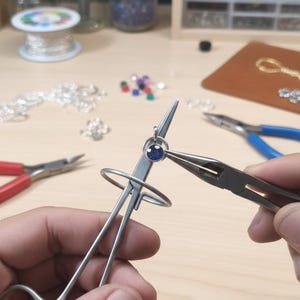 Puede incluir: Herramientas para hacer joyas en uso. Un par de alicates sostiene un pequeño anillo de plata con una piedra preciosa azul, mientras que otra herramienta se utiliza para manipular el anillo. Otros suministros para hacer joyas son visibles en el fondo.