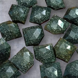 Puede incluir: Una colección de gemas facetadas de color verde oscuro con un aspecto brillante y moteado. Las piedras tienen forma irregular, con respaldos planos y múltiples facetas talladas en las superficies superiores. Las gemas están dispuestas juntas.