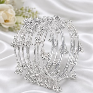 May include: A set of five silver-toned bangles featuring a textured, ridged surface. Each bangle is adorned with multiple small, dangling bell-shaped charms attached at regular intervals along the entire circumference of the circular metal bands.