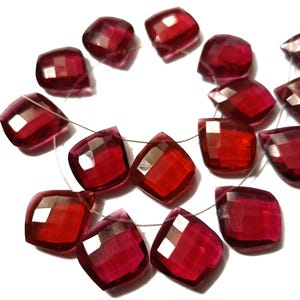 May include: A collection of faceted, square-shaped red gemstones strung together on a thin, clear filament. Each stone features a polished surface with multiple geometric facets that reflect light, creating a vibrant, translucent crimson appearance.