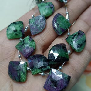 May include: A strand of faceted gemstone beads. The beads are a mix of green and purple, with a geometric cut. The beads are strung on a thin, silver-colored wire. The gemstones have a polished, reflective surface.