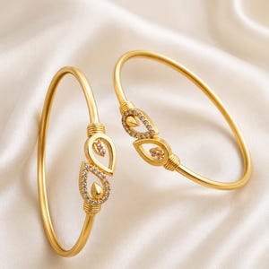 May include: Two gold-toned bangles featuring a teardrop-shaped focal point. Each teardrop is outlined with small clear stones and contains a single pink stone at the center. The bangles have a smooth, polished finish with ribbed detailing.