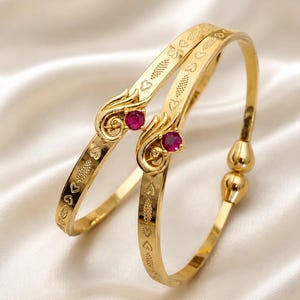 May include: Two gold-toned bangles featuring a polished finish with embossed heart and leaf patterns along the band. Each bangle is accented with a single round, vibrant red gemstone set within a decorative, swirling gold-toned metal flourish.