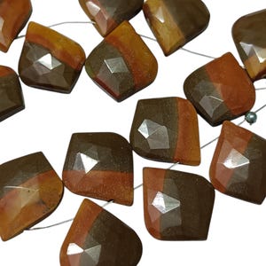 Puede incluir: Un conjunto de cuentas facetadas de piedras preciosas en tonos marrón, naranja y beige. Las cuentas están ensartadas en un fino hilo plateado. Cada cuenta tiene una forma geométrica única con múltiples facetas.