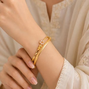 May include: A set of two gold-toned bangles featuring a delicate flower-shaped charm with small white stones. One bangle has a smooth, polished finish, while the other displays a subtle textured pattern along its surface, creating a layered look.