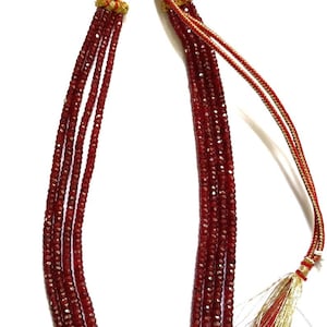 Peut inclure: Un collier multi-rangs composé de quatre rangées de perles facettées rouge foncé. Les brins sont fixés par des embouts en métal doré et reliés à un cordon tressé rouge et or. Un pompon décoratif en fils rouge et or métallique termine l'ensemble.
