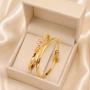 Peut inclure: Deux bracelets rigides dorés reposant dans une boîte à bijoux beige. L'un des bracelets présente une breloque en forme de fleur avec des pierres claires, tandis que l'autre affiche un motif gravé. Les deux bracelets ont une finition polie.