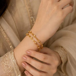 May include: Two gold bangles featuring a repeating geometric pattern with small, clear, inset stones. The bracelets have a polished metallic finish and a slender, circular design, worn together on the wrist to create a layered appearance.