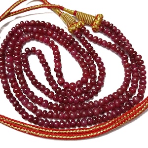 Op de afbeelding: Een meerstrengs ketting bestaande uit kleine, ronde, dieprode kralen. De ketting heeft goudkleurige accenten en rode en gouden koorden aan de uiteinden. De kralen zijn gelijkmatig van formaat en strak geregen, wat een rijke, getextureerde uitstraling creëert.