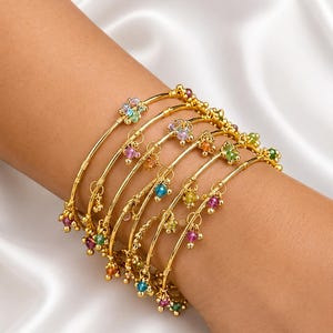 Puede incluir: Un conjunto de varias pulseras rígidas doradas usadas juntas, cada una con pequeños dijes colgantes de piedras preciosas multicolores en tonos rosa, azul, verde y amarillo. Las finas bandas de metal pulido crean un aspecto superpuesto.