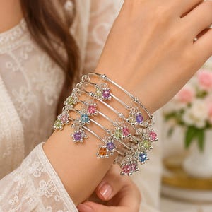 Könnte beinhalten: Ein Set aus fünf silberfarbenen Armreifen, die jeweils mit mehreren kleinen, baumelnden, facettierten, bunten Kristallanhängern in den Farben Rosa, Lila, Blau, Grün und Orange versehen und am Handgelenk gestapelt sind.
