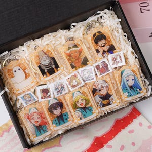 Puede incluir: Un conjunto de llaveros de personajes de estilo anime en una caja de regalo. Cada llavero presenta una ilustración de personaje diferente con un marco dorado. También se incluyen pequeñas tarjetas de personajes. La caja está llena de papel triturado blanco.