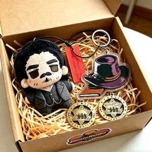 May include: A gift box filled with a plush doll, keychains, and medallions. The plush doll has a black hairstyle and a mustache. The keychains feature a hat and book design. The medallions have a star shape with the words "RED DEAD REDEMPTION".