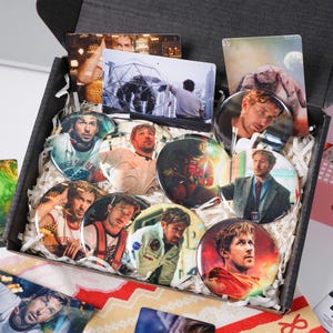 May include: A black box containing various items, including several round pin badges and rectangular cards. The badges feature images of a man in different outfits. The box is lined with white paper shreds.