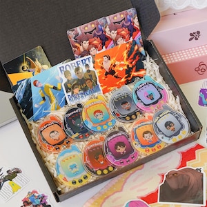 May include: A black box filled with colorful character keychains and cards. The keychains have various character designs, and the cards feature illustrations of characters. The box is lined with white paper and includes stickers.