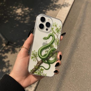 Puede incluir: Una funda transparente para smartphone con una ilustración detallada de una serpiente verde brillante enroscada alrededor de una rama de árbol marrón texturizada con pequeñas hojas verdes. El diseño está impreso en la parte trasera.
