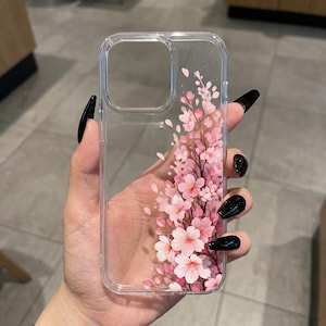 Può includere: Custodia trasparente per telefono con un motivo floreale rosa. Il design presenta fiori di ciliegio rosa e foglie, creando un'estetica primaverile. La custodia è realizzata in un materiale trasparente.