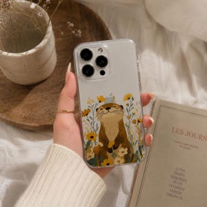 Könnte beinhalten: Eine transparente Smartphone-Hülle mit einer Illustration eines braunen Otters inmitten gelber und weißer Wildblumen. Die Hülle hat eine Aussparung für ein Triple-Kamera-System und zeigt das Apple-Logo durch das durchsichtige Material.
