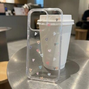 May include: Transparent phone case adorned with small, pastel-coloured hearts. The case is made of clear plastic and is designed for a smartphone. The hearts are in shades of pink, blue, and yellow. The case is next to a white paper coffee cup.