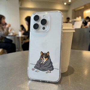 May include: Transparent phone case featuring a cartoon corgi wearing glasses and wrapped in a grey blanket. The corgi is holding a phone. The case is on a silver surface with a white coffee cup in the background.