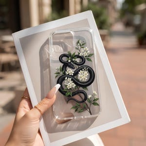 Può includere: Una custodia trasparente per telefono con un'illustrazione grafica di un serpente nero avvolto attorno a un ramo fiorito con fiori bianchi e foglie verdi, stampata sulla cover protettiva per il dispositivo mobile.