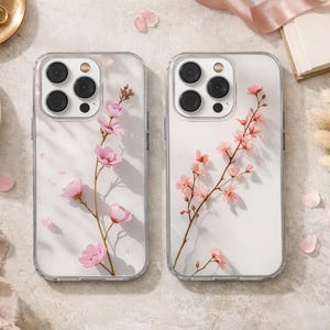 May include: Two clear phone cases with floral designs. Each case features a delicate pink cherry blossom branch with small pink petals. The cases are displayed on a neutral surface with other accessories, including a cup of coffee and a perfume bottle.