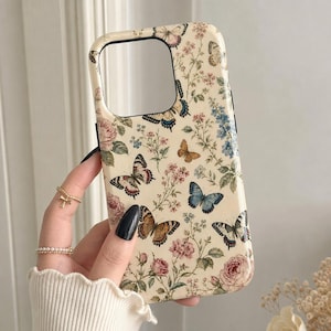 Op de afbeelding: Een crèmekleurig telefoonhoesje met een gedetailleerde botanische print met diverse vlinders in blauw-, bruin- en oranjetinten, afgewisseld met roze rozen, groene bladeren en kleine blauwe bloemen over het gehele oppervlak.