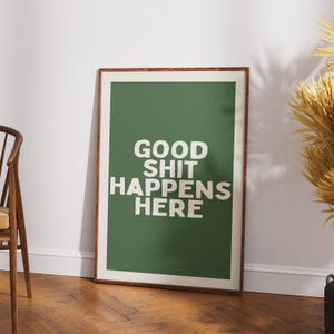 May include: A framed art print with a green background and the white text "GOOD SHIT HAPPENS HERE." The print is in a brown wooden frame and leans against a white wall on a wooden floor.