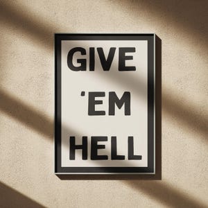 Puede incluir: Un cuadro enmarcado con la frase "GIVE 'EM HELL" en letras negras y audaces sobre un fondo crema. La impresión está encerrada en un marco negro y cuelga de una pared beige texturizada.