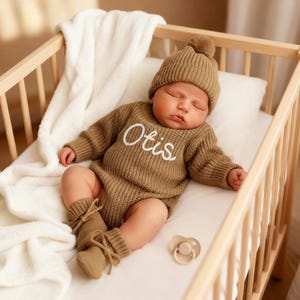 Może przedstawiać: Śpiące dziecko w brązowym dzianinowym stroju, w tym czapka z pomponem, sweter z napisem "Otis" i pasujące skarpetki. Dziecko leży w drewnianym łóżeczku z białym kocem i poduszką oraz smoczkiem.