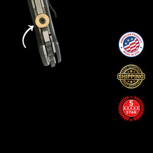 Pode incluir: Close-up de uma faca dobr&aacute;vel parcialmente vis&iacute;vel com cabo preto e l&acirc;mina prateada. Um parafuso dourado &eacute; vis&iacute;vel na lateral da faca. A imagem tamb&eacute;m inclui um emblema "Made in USA" e um emblema "5 estrelas".