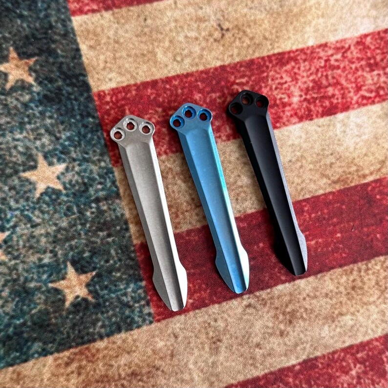 May include: Three metal knife scales in silver, blue, and black, each with a three-hole design at the top. The scales are displayed on a backdrop of the American flag, with red and white stripes and a blue field of stars.