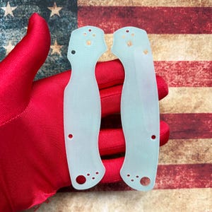 May include: Two light blue knife handle scales with multiple holes and a textured surface are displayed against a backdrop of the American flag. The scales are held by a red gloved hand.