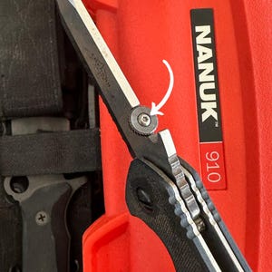 Puede incluir: Primer plano de una navaja plegable negra y plateada con mango texturizado. La hoja est&aacute; parcialmente abierta, mostrando un tornillo de pivote plateado. El cuchillo est&aacute; sobre un fondo rojo con el texto "NANUK 910".
