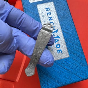 Op de afbeelding: Een zilverkleurige zakmesclip met een gegraveerd visschubbenpatroon. De clip bevindt zich boven een blauwe doos met de tekst "BENCHMADE KNIFE". De achtergrond is rood.