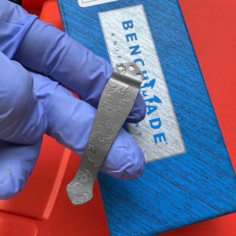 Op de afbeelding: Een zilverkleurige zakmesclip met een gegraveerd visschubbenpatroon. De clip bevindt zich boven een blauwe doos met de tekst "BENCHMADE KNIFE". De achtergrond is rood.
