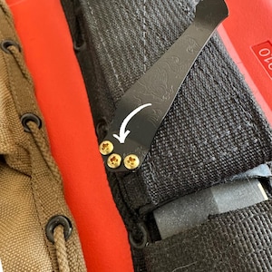 May include: Close-up of a black knife sheath with a decorative metal clip. The clip is attached with three gold-colored screws and features an etched design. A white arrow indicates the direction of the clip's opening. The sheath is made of black fabric.