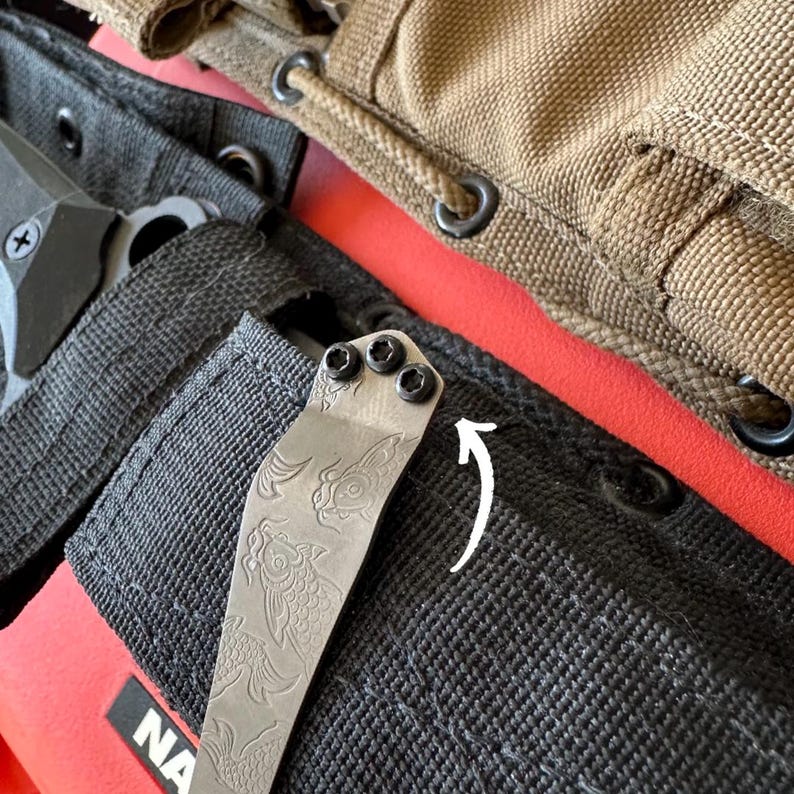 May include: Close-up of a knife clip with a fish design, attached to a black sheath. The clip is secured with three black screws. A tan sheath and a red surface are in the background.