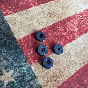 Pu&ograve; includere: Quattro piccoli oggetti circolari blu scuro con un foro centrale e una superficie a motivi. Gli oggetti sono sparsi su uno sfondo a strisce rosse, bianche e blu, che ricorda la bandiera americana.