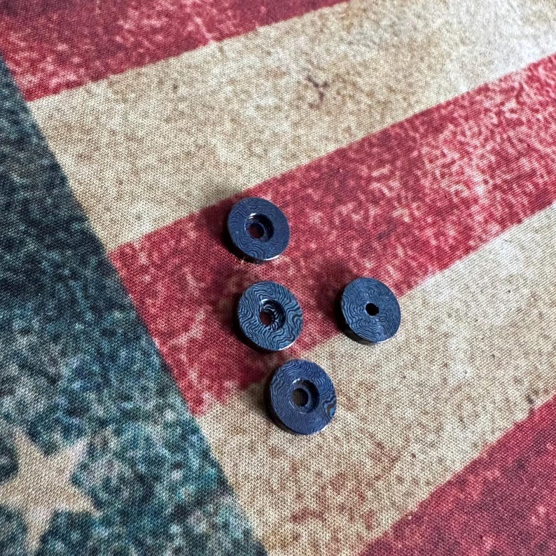 Pu&ograve; includere: Quattro piccoli oggetti circolari blu scuro con un foro centrale e una superficie a motivi. Gli oggetti sono sparsi su uno sfondo a strisce rosse, bianche e blu, che ricorda la bandiera americana.