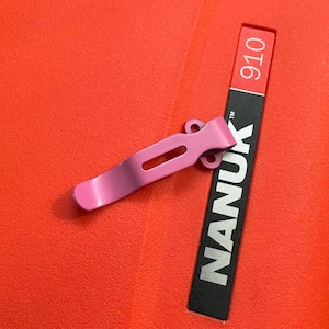 May include: A pink metal clip with a curved design and a rectangular cutout, resting on a bright red surface. A black label with the word "NANUK" in white and the number "910" is also visible.