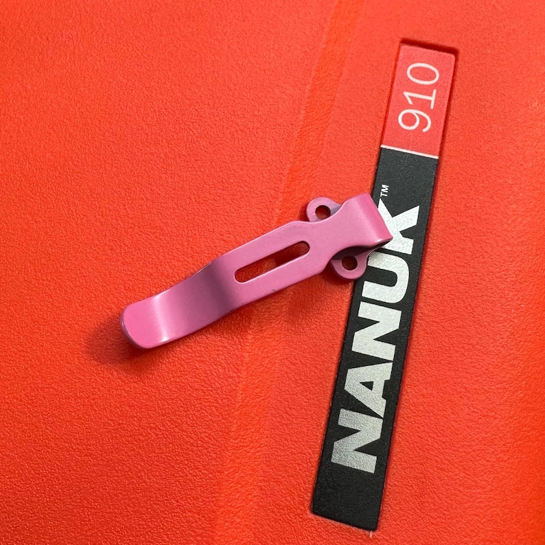 May include: A pink metal clip with a curved design and a rectangular cutout, resting on a bright red surface. A black label with the word "NANUK" in white and the number "910" is also visible.