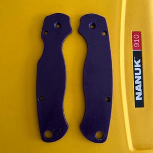 May include: Two purple knife handle scales with multiple holes for attachment. The scales are made of a textured material and are shown against a yellow background. The text "NANUK 910" is visible on the right side.