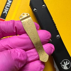 May include: A gold-colored metal pocket clip featuring an engraved koi fish pattern. The clip has three circular mounting holes at the top and a curved shape. It is positioned next to a black handle with silver screws and a circular logo.