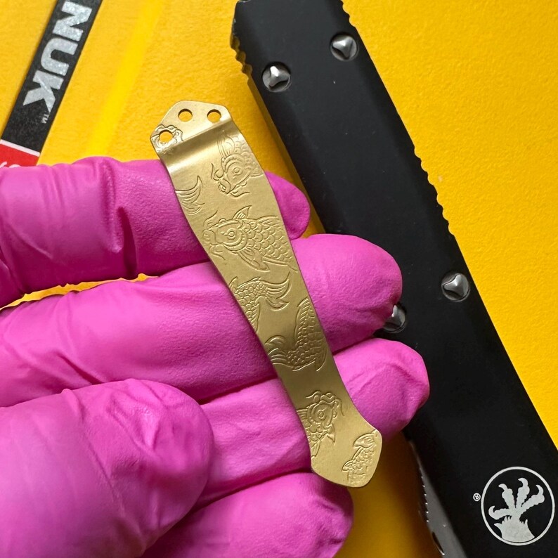 May include: A gold-colored metal pocket clip featuring an engraved koi fish pattern. The clip has three circular mounting holes at the top and a curved shape. It is positioned next to a black handle with silver screws and a circular logo.